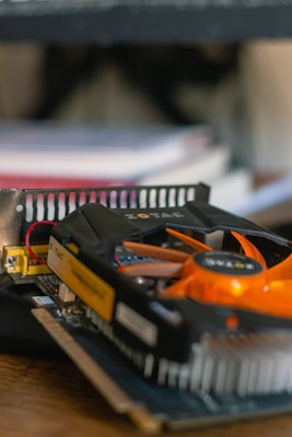 A close-up view of a computer graphics card with a prominent orange fan and black housing. The card is connected to a motherboard and surrounded by other computer components. In the background, there are blurred books or documents, implying a workspace setting.