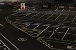 A dimly lit empty parking lot with marked parking spaces including numbered spots and painted arrows. There is a security booth at one end, surrounded by traffic cones and a gate mechanism. The area is fenced in and appears to have been photographed at night.