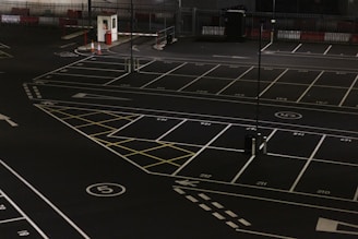 A dimly lit empty parking lot with marked parking spaces including numbered spots and painted arrows. There is a security booth at one end, surrounded by traffic cones and a gate mechanism. The area is fenced in and appears to have been photographed at night.