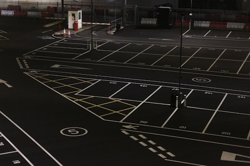 A dimly lit empty parking lot with marked parking spaces including numbered spots and painted arrows. There is a security booth at one end, surrounded by traffic cones and a gate mechanism. The area is fenced in and appears to have been photographed at night.