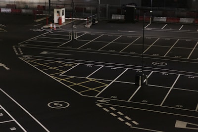 A dimly lit empty parking lot with marked parking spaces including numbered spots and painted arrows. There is a security booth at one end, surrounded by traffic cones and a gate mechanism. The area is fenced in and appears to have been photographed at night.