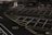 A dimly lit empty parking lot with marked parking spaces including numbered spots and painted arrows. There is a security booth at one end, surrounded by traffic cones and a gate mechanism. The area is fenced in and appears to have been photographed at night.