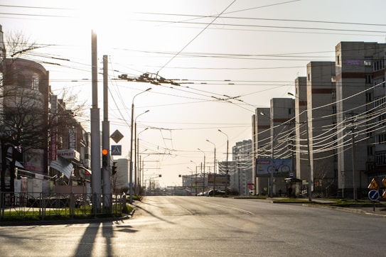 A quiet urban street scene featuring tall apartment buildings on either side. Numerous power lines crisscross the sky, illuminated by a low sun. Traffic lights and signs are visible, and the road appears empty, suggesting early morning or late evening.