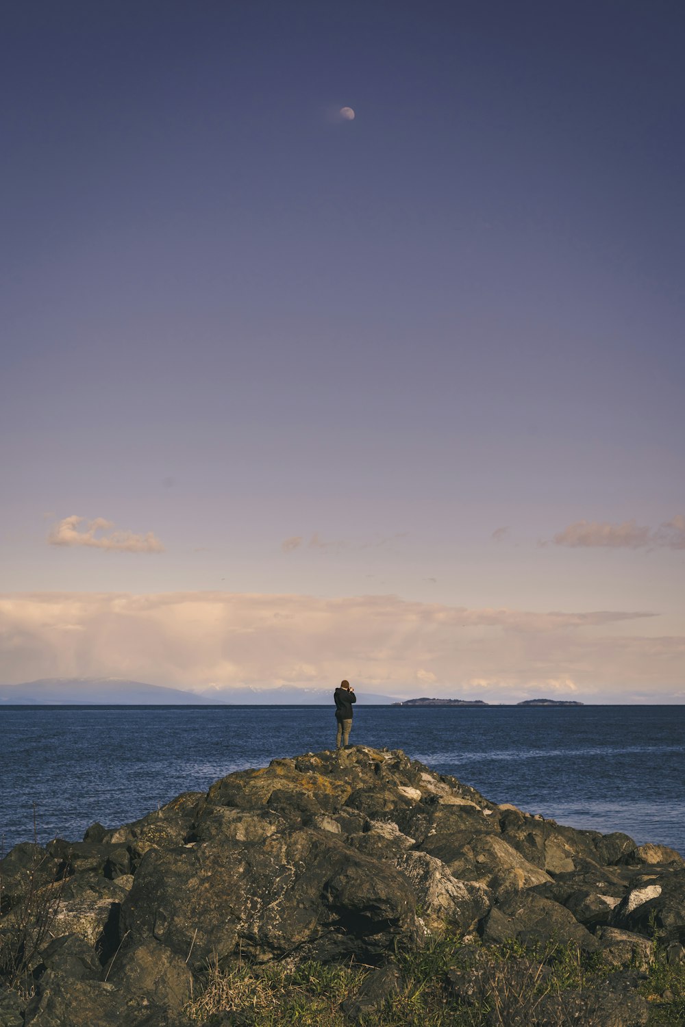 Person, die tagsüber auf einer Felsformation in der Nähe des Meeres unter blauem Himmel steht