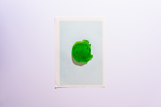 A green gel-like substance is placed on a sheet of graph paper. The paper has a white border, and the graph appears to be light blue. The gel has an irregular shape, with small indentations and a glossy surface.