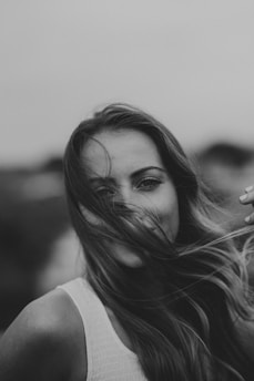 A black and white portrait of a person smiling with the wind blowing their hair across their face. The individual is wearing a textured sleeveless top, and the background is a soft blur, which gives an intimate and candid feel to the photograph.