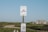 A clear blue sky over a bus stop sign for Martha's Vineyard Transit Authority is visible. The sign is mounted on a wooden post with a schedule attached below. In the background, there is a small house and green shrubbery set in a landscape that appears rural.