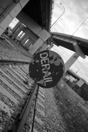 A black and white image featuring a railway track with a circular metal sign prominently displaying the word 'DERAIL'. The railway track is set beneath an overpass or bridge structure, with tall supporting pillars in the background. The setting appears urban and industrial, with scattered gravel and metal components.