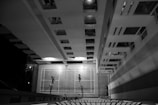 A top-down black and white view of a building with a badminton court located in the courtyard. Three people are actively playing, casting long shadows across the court, while the surrounding windows and structure of the building create a geometric pattern.