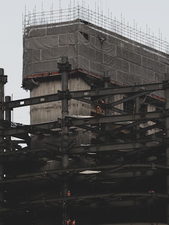 A building under construction with numerous steel beams and scaffolding. The upper section is covered with a protective fabric, and workers in bright clothing are visible on the structure, indicating ongoing construction activity.