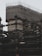A building under construction with numerous steel beams and scaffolding. The upper section is covered with a protective fabric, and workers in bright clothing are visible on the structure, indicating ongoing construction activity.