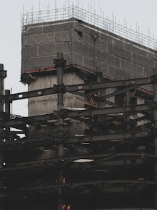 A building under construction with numerous steel beams and scaffolding. The upper section is covered with a protective fabric, and workers in bright clothing are visible on the structure, indicating ongoing construction activity.