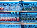 Shelves in a store are stocked with multiple brands of bottled water, neatly arranged with clear plastic bottles and colorful labels. Price tags are visible on the shelves.