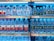 Shelves in a store are stocked with multiple brands of bottled water, neatly arranged with clear plastic bottles and colorful labels. Price tags are visible on the shelves.