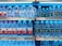 Shelves in a store are stocked with multiple brands of bottled water, neatly arranged with clear plastic bottles and colorful labels. Price tags are visible on the shelves.