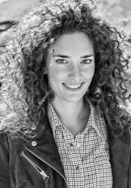 A black and white portrait of a person with curly hair, smiling pleasantly. The individual is wearing a checked shirt and a jacket with zipper detail. The background appears to be out of focus, adding emphasis to the subject's facial expression.