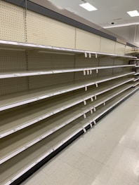 Empty supermarket shelves with product tags attached. The shelves are uniformly lined and the lighting suggests a bright indoor setting.