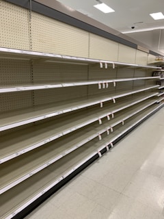 Empty supermarket shelves with product tags attached. The shelves are uniformly lined and the lighting suggests a bright indoor setting.