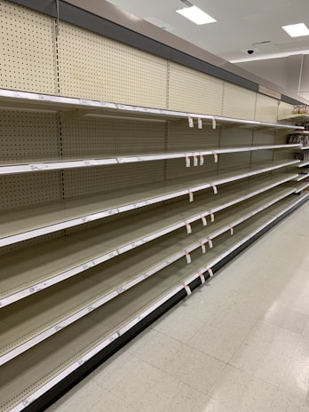 Empty supermarket shelves with product tags attached. The shelves are uniformly lined and the lighting suggests a bright indoor setting.