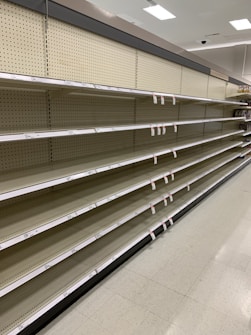 Empty supermarket shelves with product tags attached. The shelves are uniformly lined and the lighting suggests a bright indoor setting.