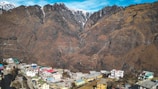 A small town is nestled at the base of towering, rugged mountains. The town features colorful buildings with vibrant rooftops. The mountains have a stark, rocky appearance with patches of snow near the peaks and are set against a bright blue sky.
