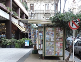 A small kiosk covered with various newspapers and magazines is situated on a sidewalk. The kiosk is surrounded by urban buildings and greenery, with a no-right-turn traffic sign visible nearby. Pedestrians can be seen in the background browsing other shops.
