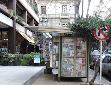 A small kiosk covered with various newspapers and magazines is situated on a sidewalk. The kiosk is surrounded by urban buildings and greenery, with a no-right-turn traffic sign visible nearby. Pedestrians can be seen in the background browsing other shops.
