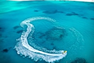 A jet ski creates a swirling pattern on the clear, turquoise ocean water near a sandy beach. The wake forms a prominent curved trail as the jet ski moves swiftly across the surface.