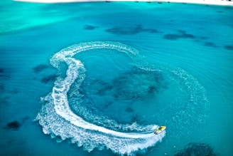 A jet ski creates a swirling pattern on the clear, turquoise ocean water near a sandy beach. The wake forms a prominent curved trail as the jet ski moves swiftly across the surface.
