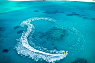 A jet ski creates a swirling pattern on the clear, turquoise ocean water near a sandy beach. The wake forms a prominent curved trail as the jet ski moves swiftly across the surface.