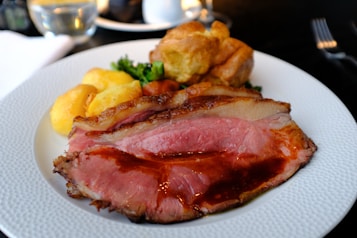 A plate of roasted meat with a slice of beef cooked to medium rare, accompanied by roasted golden potatoes and a Yorkshire pudding. The meal is garnished with some greens, possibly parsley or leafy vegetables, and is presented on a white plate with a textured rim. The background includes a softly lit setting with a glass of water and a fork partially visible.