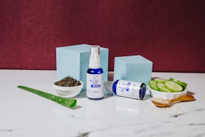A blue and white bottle of night serum stands upright next to a matching bottle lying on its side. These are placed on a white marble surface, surrounded by natural ingredients like sliced cucumbers, a piece of aloe vera, a small bowl of tea leaves, and a wooden spoon filled with a granular substance. Two light blue boxes add a geometric element in the background, against a deep burgundy wall.