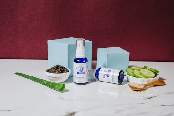 A blue and white bottle of night serum stands upright next to a matching bottle lying on its side. These are placed on a white marble surface, surrounded by natural ingredients like sliced cucumbers, a piece of aloe vera, a small bowl of tea leaves, and a wooden spoon filled with a granular substance. Two light blue boxes add a geometric element in the background, against a deep burgundy wall.
