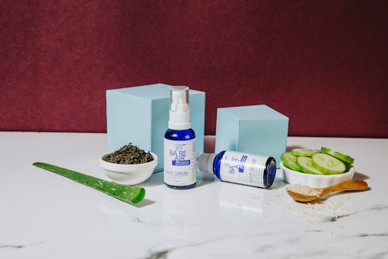 A blue and white bottle of night serum stands upright next to a matching bottle lying on its side. These are placed on a white marble surface, surrounded by natural ingredients like sliced cucumbers, a piece of aloe vera, a small bowl of tea leaves, and a wooden spoon filled with a granular substance. Two light blue boxes add a geometric element in the background, against a deep burgundy wall.