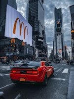 A vibrant urban street scene features a bright red sports car driving through a bustling city center. Tall skyscrapers are adorned with large digital advertisements, including a prominent McDonald's logo. The sky is cloudy, adding a moody tone to the busy atmosphere.