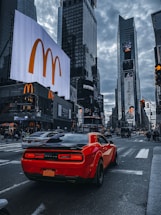 A vibrant urban street scene features a bright red sports car driving through a bustling city center. Tall skyscrapers are adorned with large digital advertisements, including a prominent McDonald's logo. The sky is cloudy, adding a moody tone to the busy atmosphere.