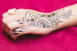 An intricately designed henna pattern is applied on the back of a hand, featuring floral and geometric motifs. The hand also wears a gold ring with a white stone, set against a rich maroon fabric background.