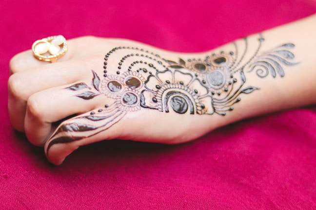 An intricately designed henna pattern is applied on the back of a hand, featuring floral and geometric motifs. The hand also wears a gold ring with a white stone, set against a rich maroon fabric background.
