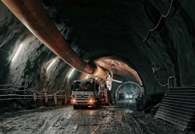 The image depicts an underground tunnel with a large vehicle, possibly a cement truck or mining equipment, positioned inside. The tunnel walls are lined with a series of evenly spaced lights providing illumination. A large ventilation duct runs along the ceiling. The tunnel floor appears muddy and wet, suggesting an industrial or construction environment.