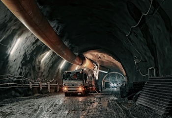 The image depicts an underground tunnel with a large vehicle, possibly a cement truck or mining equipment, positioned inside. The tunnel walls are lined with a series of evenly spaced lights providing illumination. A large ventilation duct runs along the ceiling. The tunnel floor appears muddy and wet, suggesting an industrial or construction environment.