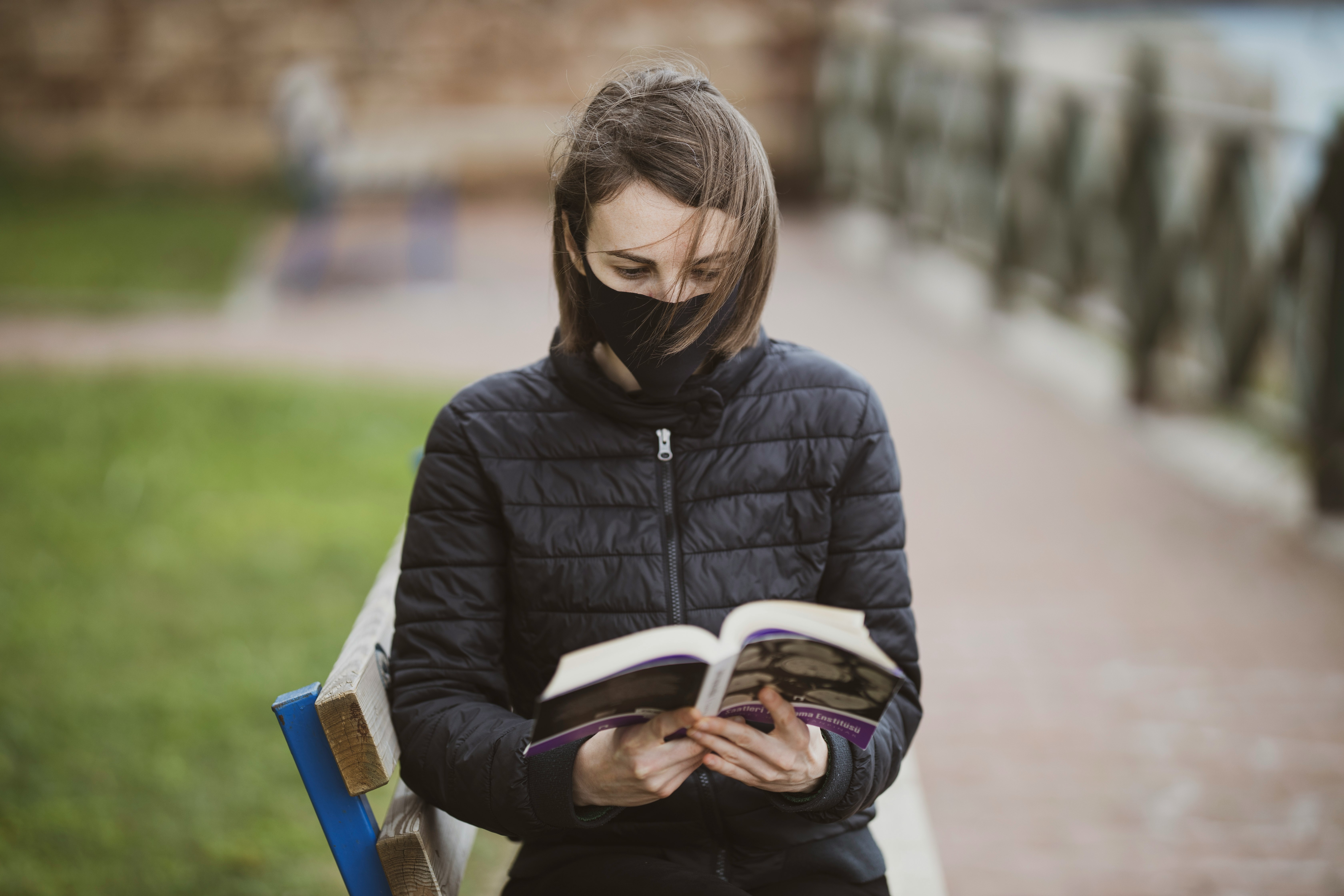 woman in black jacket sitting on bench while reading book during daytime