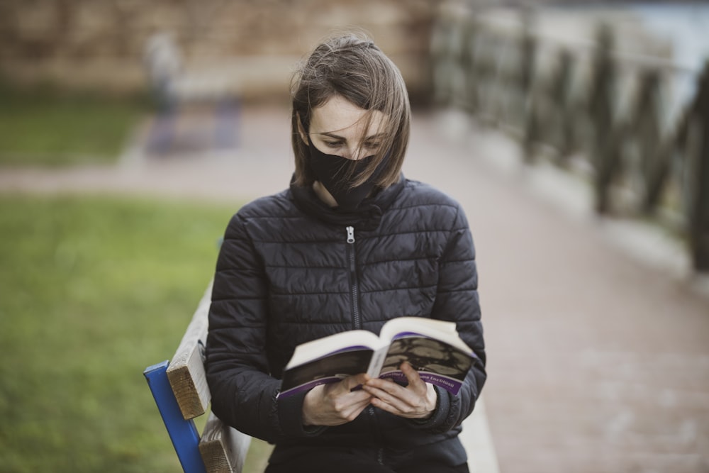 woman in black jacket sitting on bench while reading book during daytime