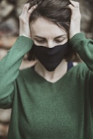 A person wearing a black mask and green sweater is holding their head with both hands. The background is blurred, focusing on the upper half of the person's body. Their eyes are closed, and they seem to be in a moment of contemplation or distress.