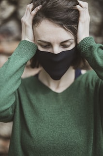 A person wearing a black mask and green sweater is holding their head with both hands. The background is blurred, focusing on the upper half of the person's body. Their eyes are closed, and they seem to be in a moment of contemplation or distress.