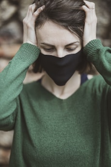 A person wearing a black mask and green sweater is holding their head with both hands. The background is blurred, focusing on the upper half of the person's body. Their eyes are closed, and they seem to be in a moment of contemplation or distress.