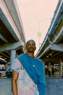 A person stands beneath an overpass, wearing a tie-dye shirt with a blue garment draped over their shoulder. The environment is urban, with concrete structures and a few other people visible in the background.