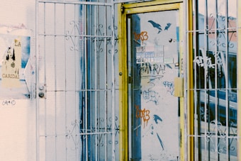 A metal gate with intricate patterns surrounds a glass door decorated with various graffiti in bright colors. The door has a yellow frame, and the glass is filled with stickers and spray-painted words. A poster with a stylized image of a girl can be seen on the adjacent wall.