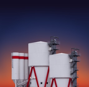 A set of large industrial silos with a modern design, featuring cylindrical white structures supported by red metal beams and external staircases. The background reveals a gradient sky transitioning from deep blue to orange, suggesting a sunset or sunrise.