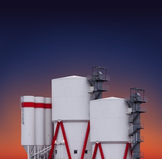 A set of large industrial silos with a modern design, featuring cylindrical white structures supported by red metal beams and external staircases. The background reveals a gradient sky transitioning from deep blue to orange, suggesting a sunset or sunrise.