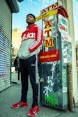 A person stands confidently next to an ATM booth covered in graffiti and stickers. They wear a red and gray Adidas hoodie, black Adidas pants, and red sneakers. The backdrop includes an urban street with a building and a partially closed metal shutter.
